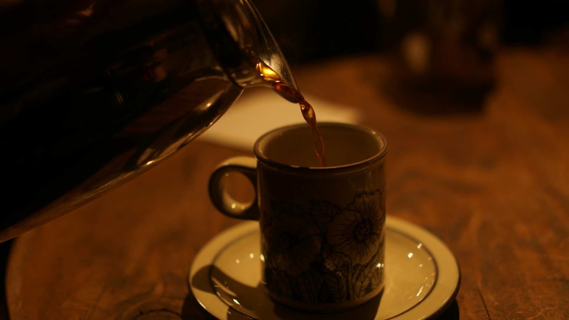 Coffee pouring from a glass carafe into a flowered cup on a saucer, in warm low light.
