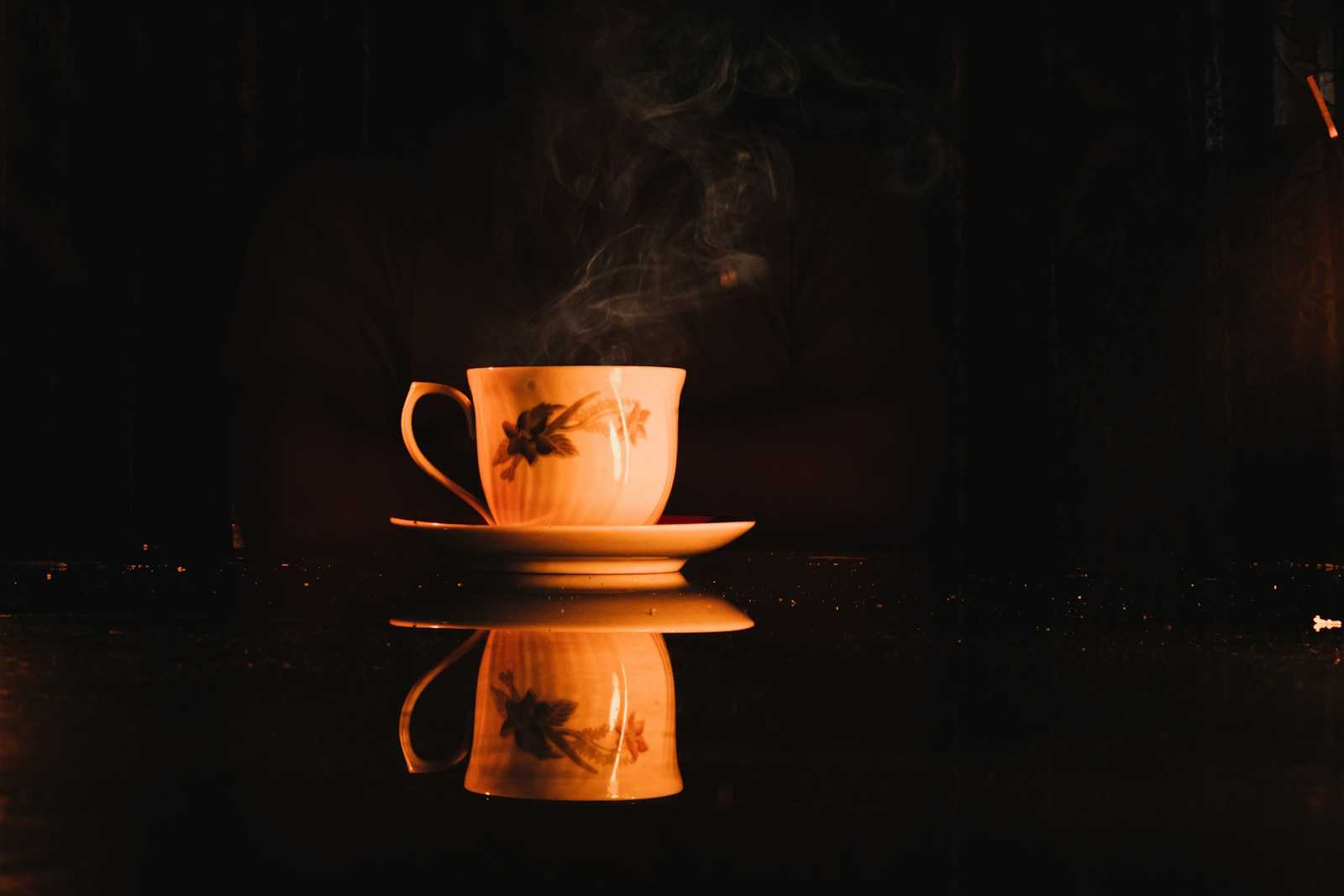 A steaming cup on a saucer, glowing amber against a dark background and reflected in the table.